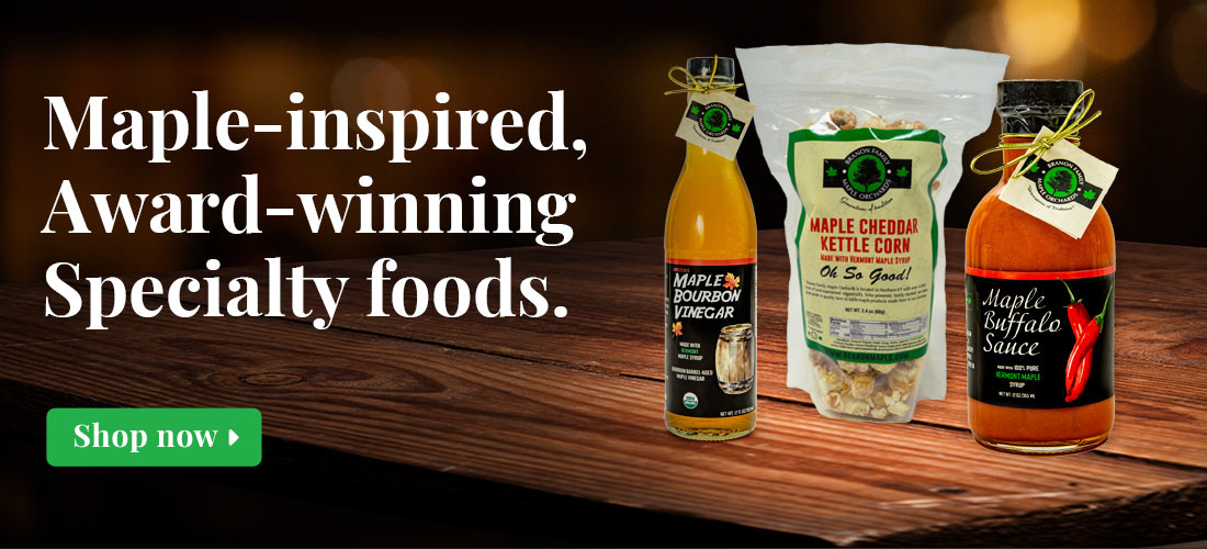 Graphic showing white serif type with green button over photo of wooden table with Organic Maple Bourbon Vinegar bottle, Maple Cheddar Kettle Corn, and Maple Buffalo Sauce